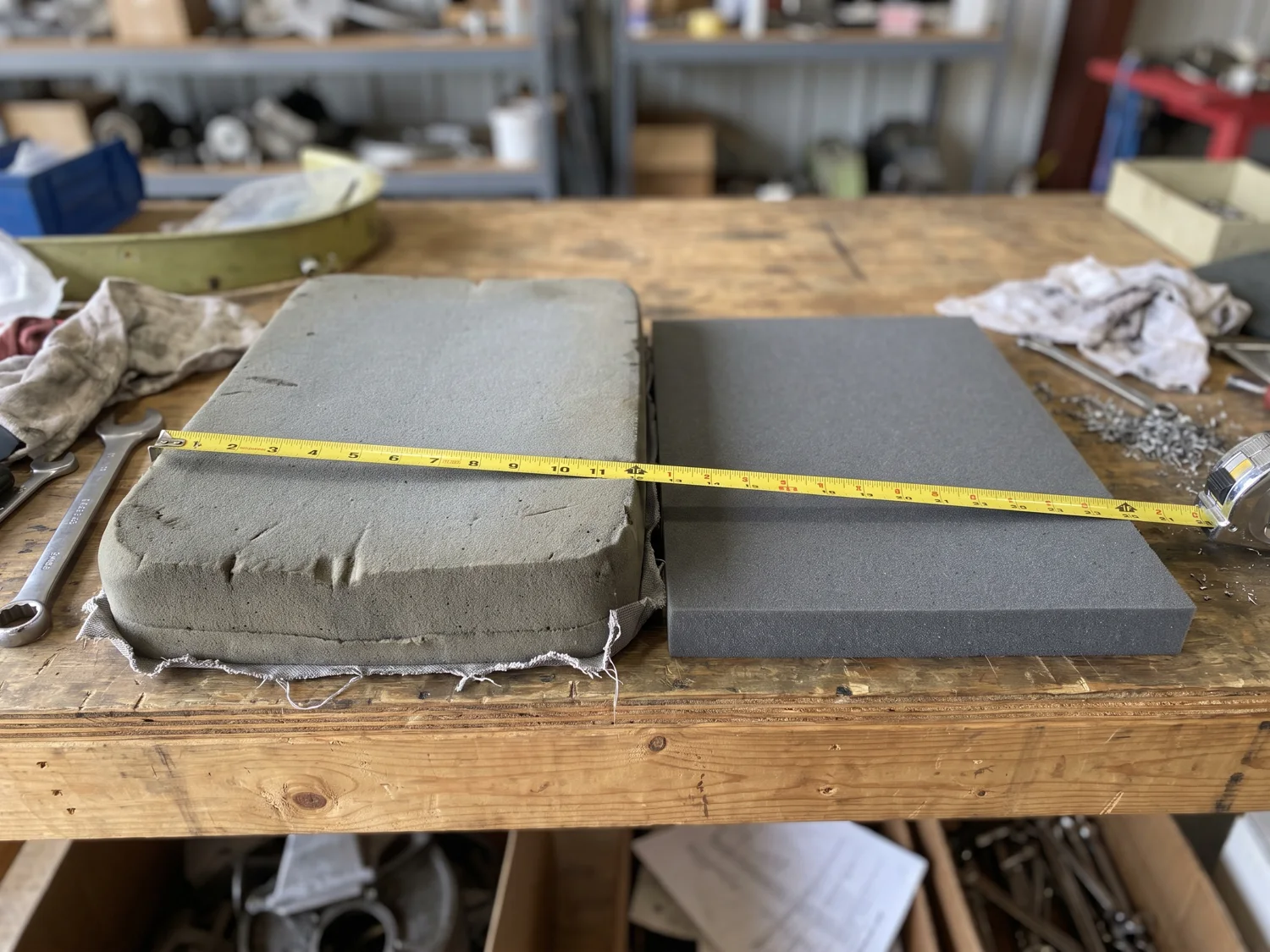Two economy seat cushions side by side on a workshop bench: the old thick one and the new one half as thick, a tape measure across them